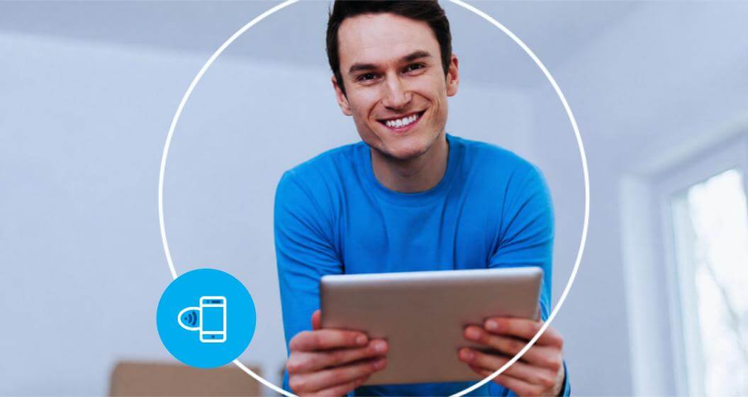 Um homem sorridente de camiseta azul segura um tablet na horizontal, emoldurado por uma moldura circular branca com um ícone de celular em destaque.O fundo apresenta um ambiente interno em tons claros e neutros, iluminado pela luz natural de uma janela.