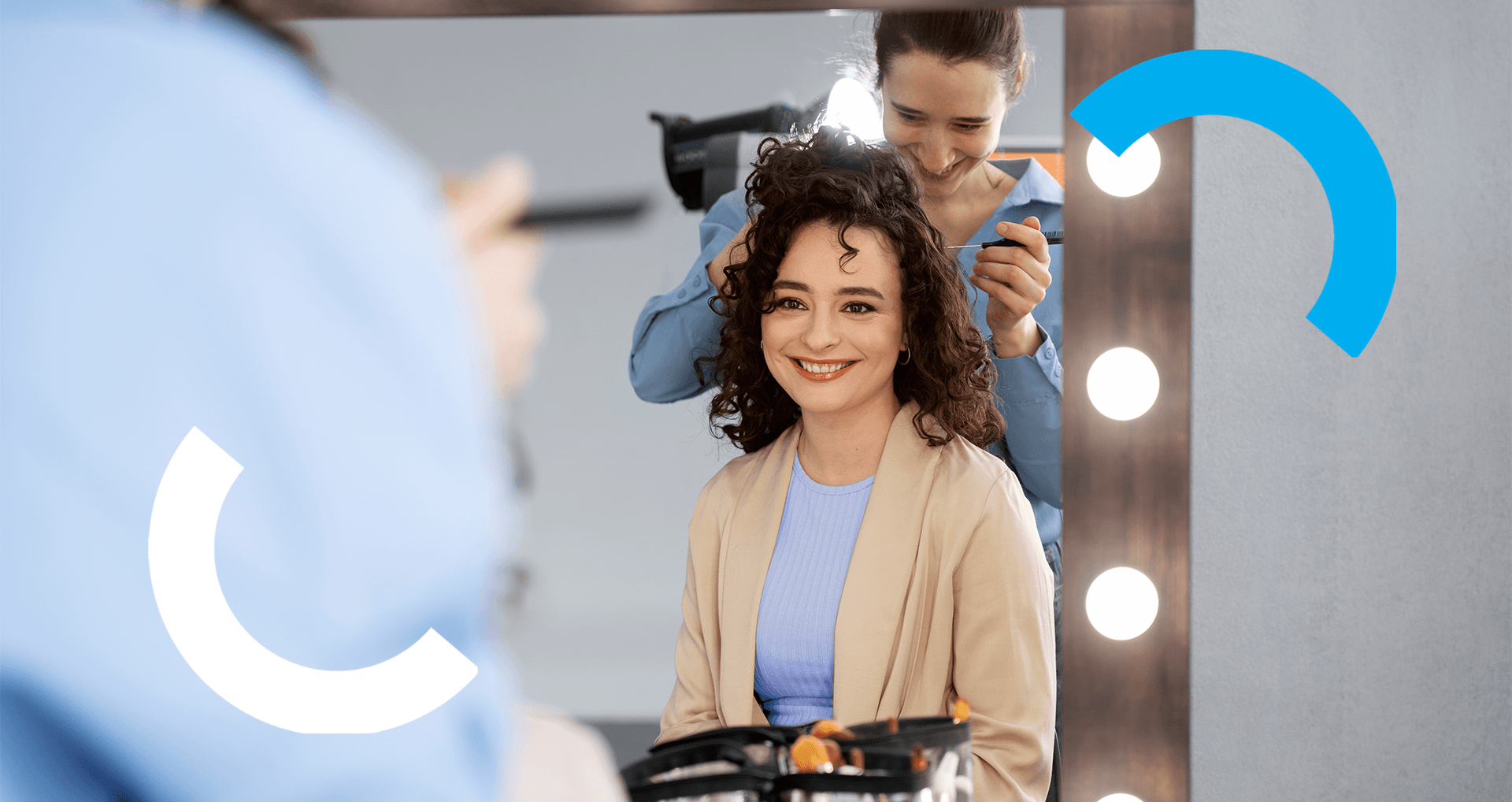 Mulher de cabelos cacheados sorri em frente ao espelho enquanto segura um smartphone, acompanhada por uma profissional ajustando seu penteado. A cena tem um espelho iluminado e grafismos semicirculares nas cores branca e azul sobre a imagem.