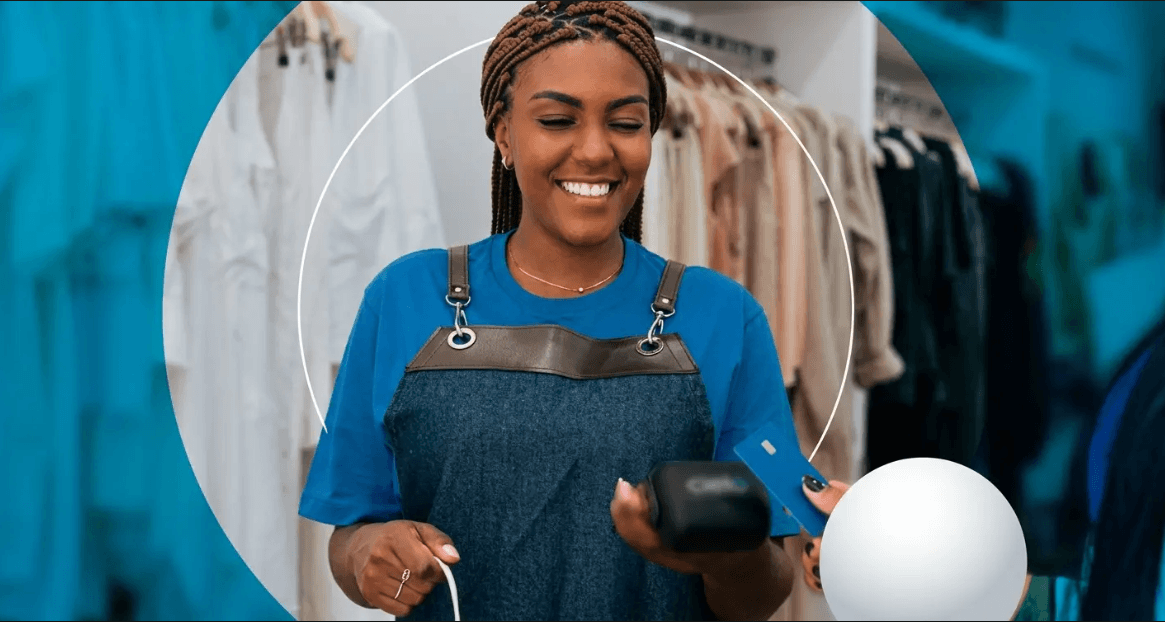 Mulher negra sorridente com tranças segura uma maquininha de cartão, enquanto outra pessoa aproxima um cartão azul para pagamento. Ela veste camiseta azul e avental de jeans, com cabides de roupas ao fundo e enquadramento circular sobre fundo azul.